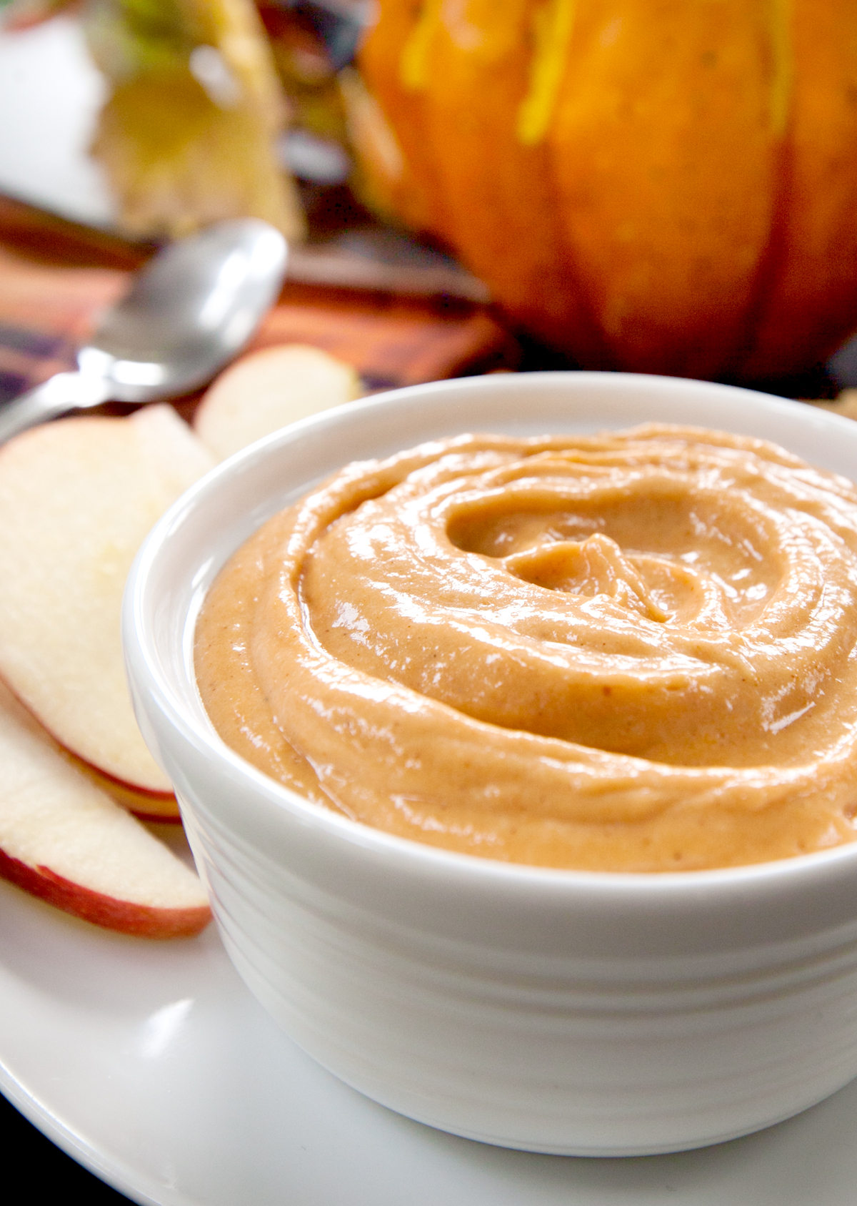 A close up of a bowl of cream cheese pumpkin dip with apple slices seen in the background for dipping.