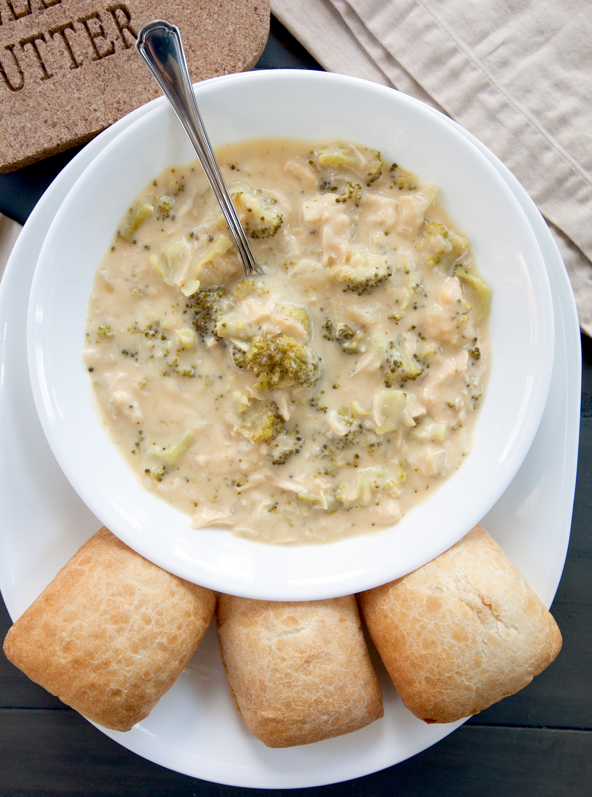 An overhead view of a bowl of chicken broccoli cheddar soup with crusty dinner rolls.