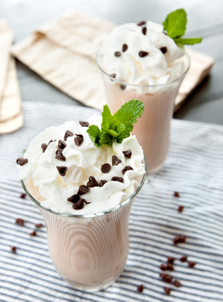 Two glasses of mint chocolate chip smoothie topped with whipped cream, mini chocolate chips, and a sprig of mint.