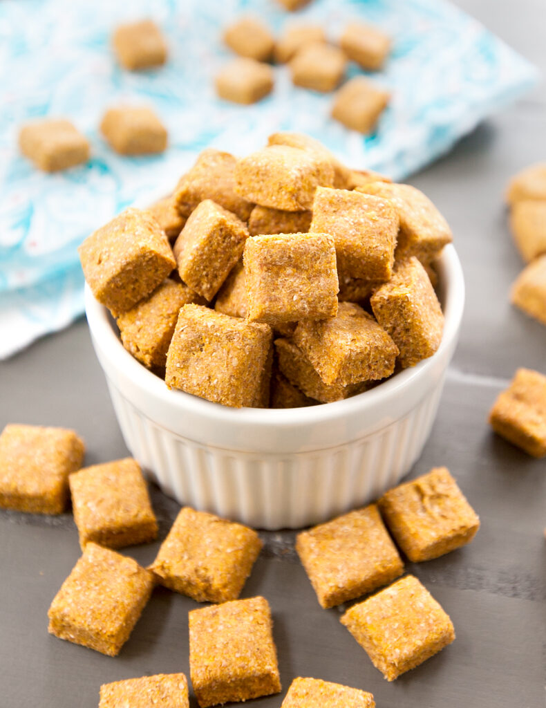 A ramekin piled high with square pumpkin banana dog treats with some scattered on the table.