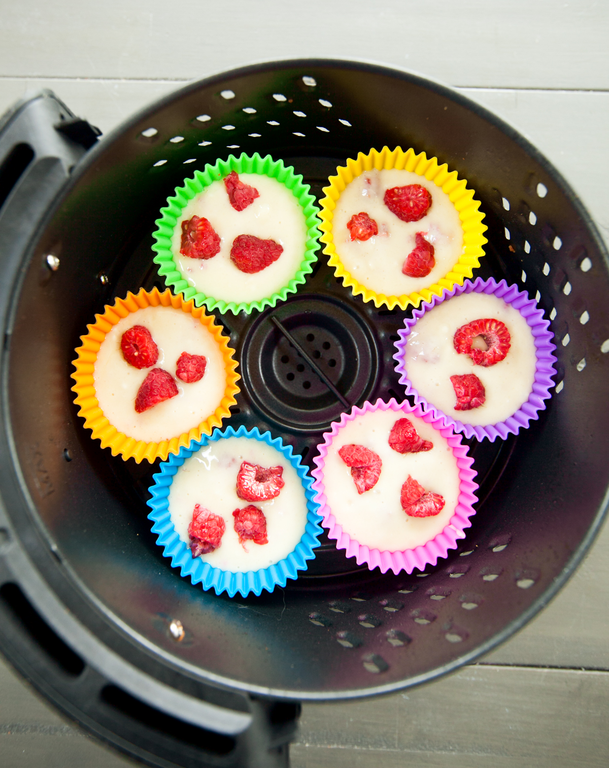 An overhead view of an air fryer basket with rainbow colored silicone baking cups filled with raspberry muffin batter.