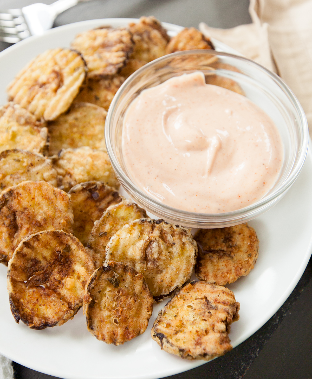 An overhead view of a bowl of dipping sauce for fried pickles surrounded by golden brown fried pickle chips.