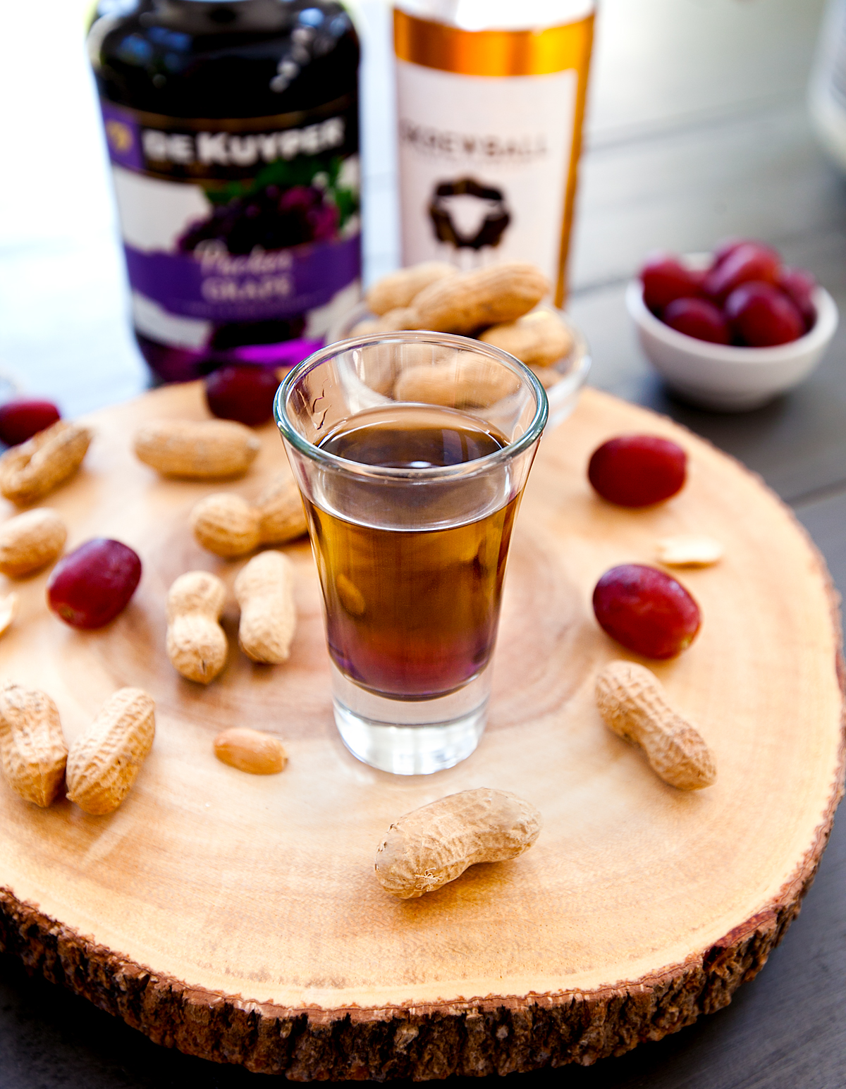 A peanut butter and jelly shot sitting on top of a slice of wood with grapes and peanuts in the shell.