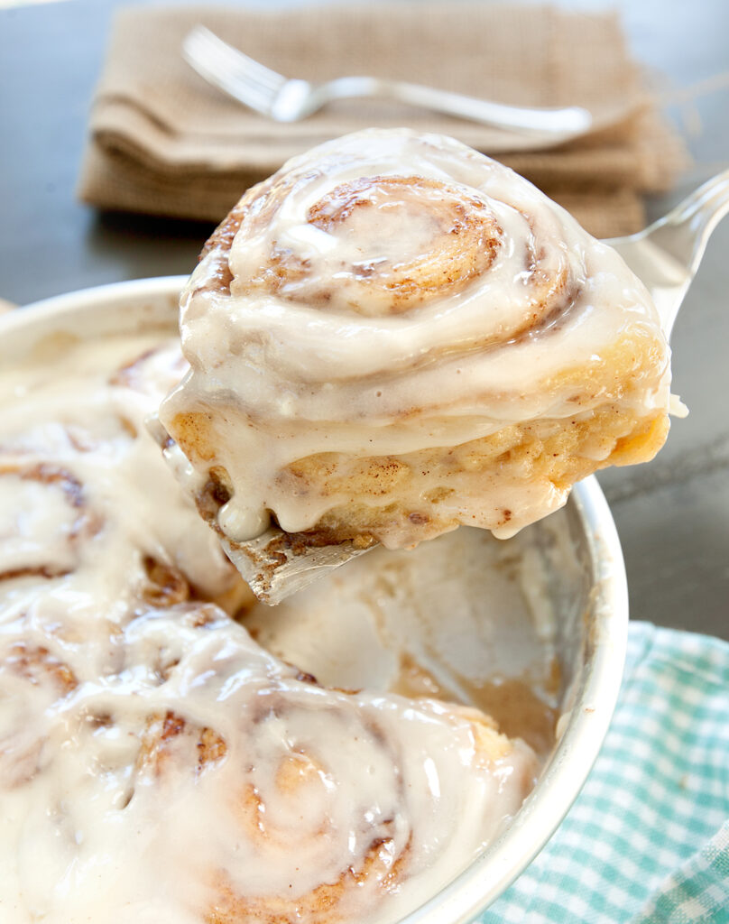 A gooey tik tok cinnamon roll is lifted out of the pan, showing plenty of frosting drizzling down.