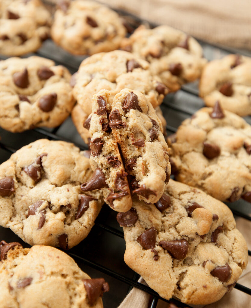 A cooling rack of oat flour chocolate chip cookies with one broken open and the halves place together so you can see all of the yummy stuff inside.