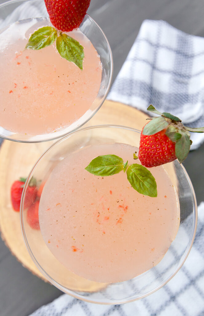 Overhead view of a pale pink strawberry martini with bits of fruit visible, decorated with a whole strawberry and floating basil leaves.