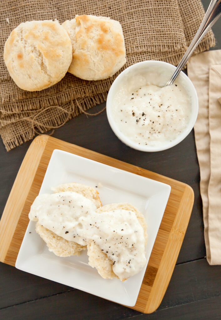 Overhead shot of biscuits and southern white gravy on a small wooden board, sprinkled with pepper.