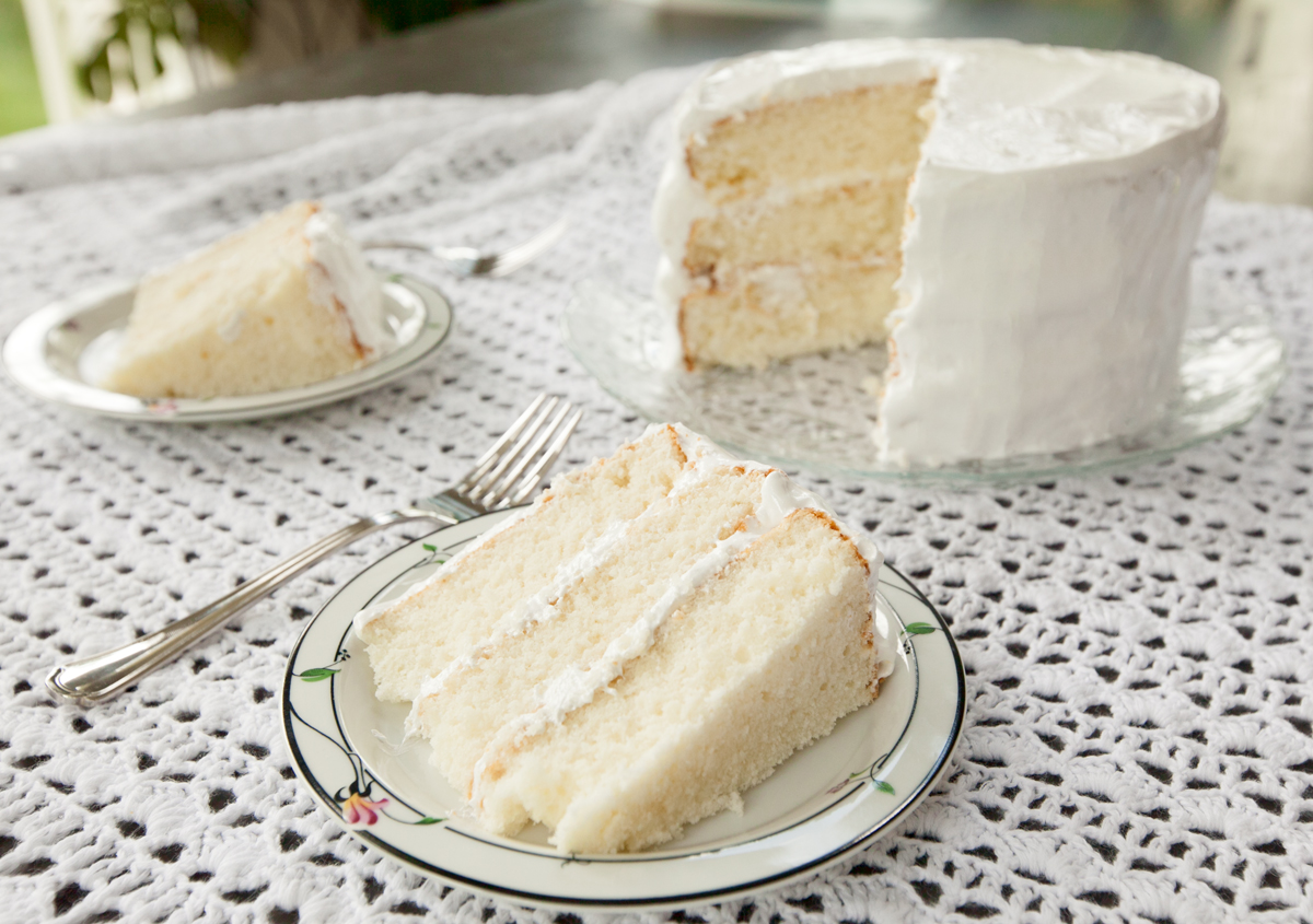 A fluffy slice of triple layer white cake with boiled frosting - inspired by Django Unchained -  sits on an old-fashioned plate over a lace tablecloth.